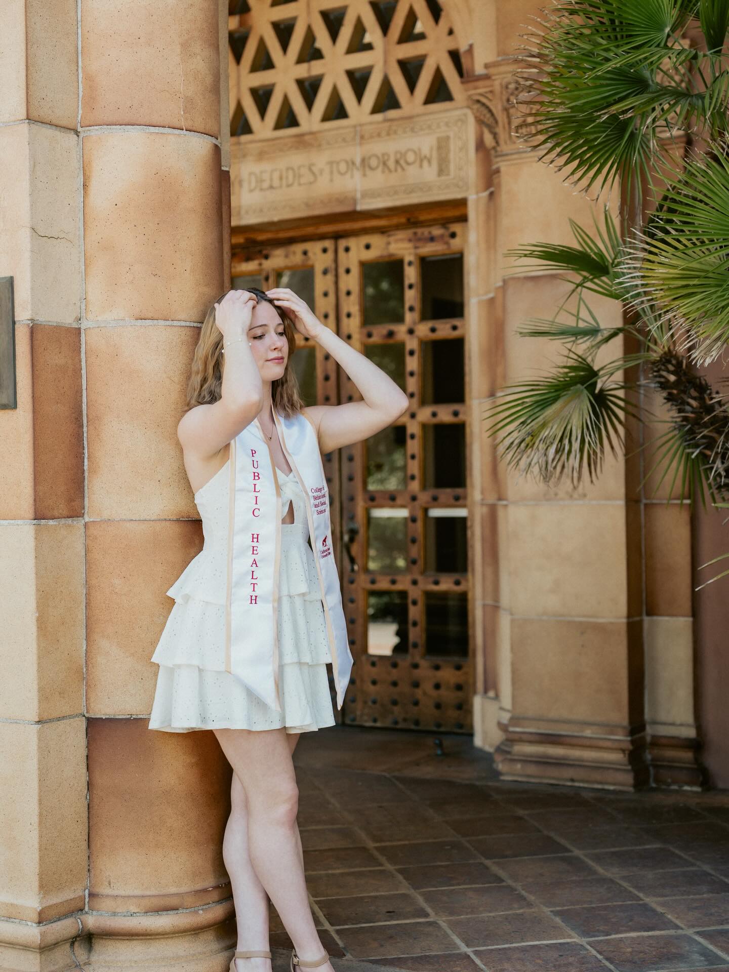 Graduate leaning against a sandstone pillar beside ornate carved doors, one hand in their hair