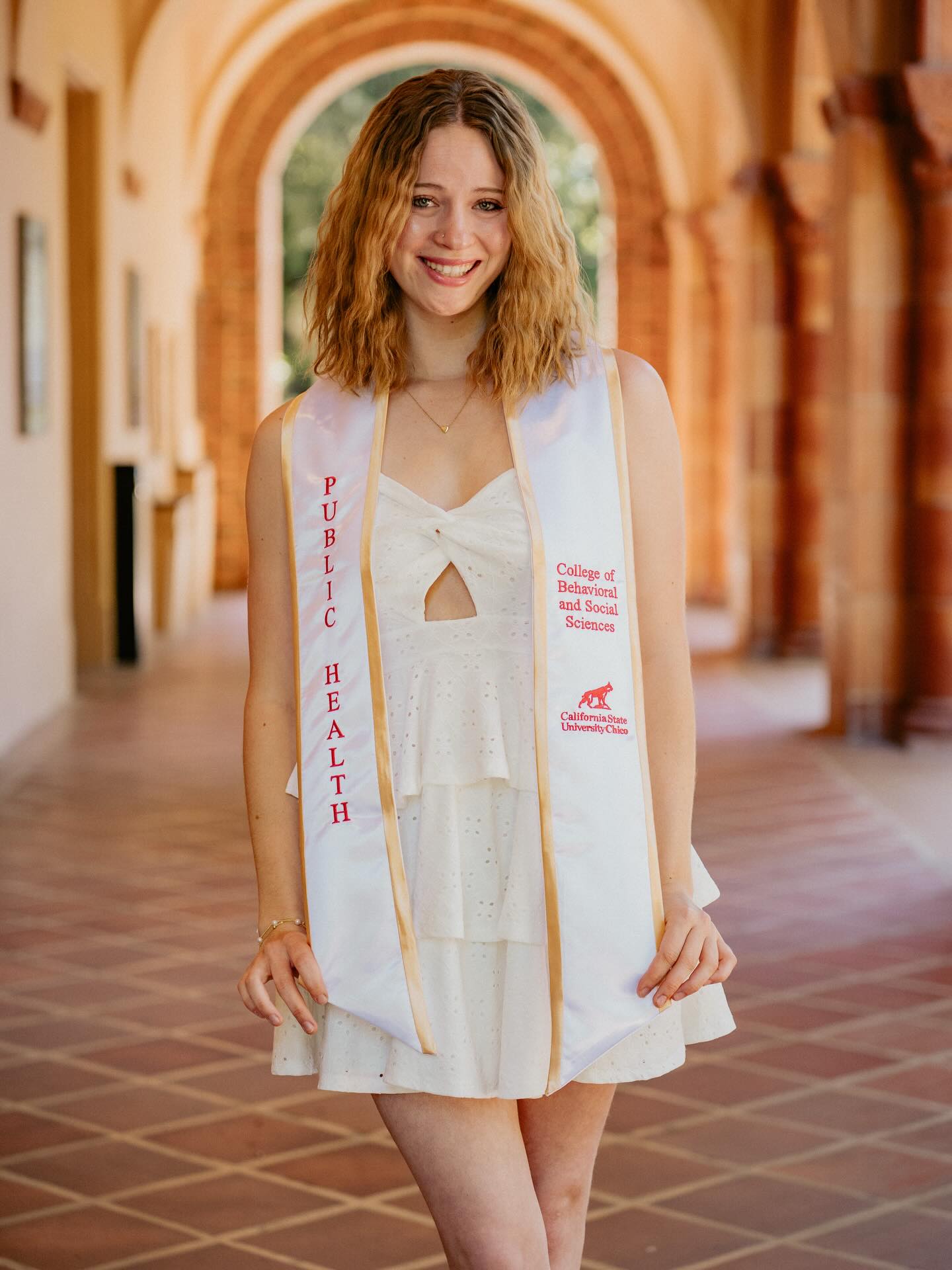 Graduate in a white dress and Public Health stole smiling in a long terracotta-arched walkway