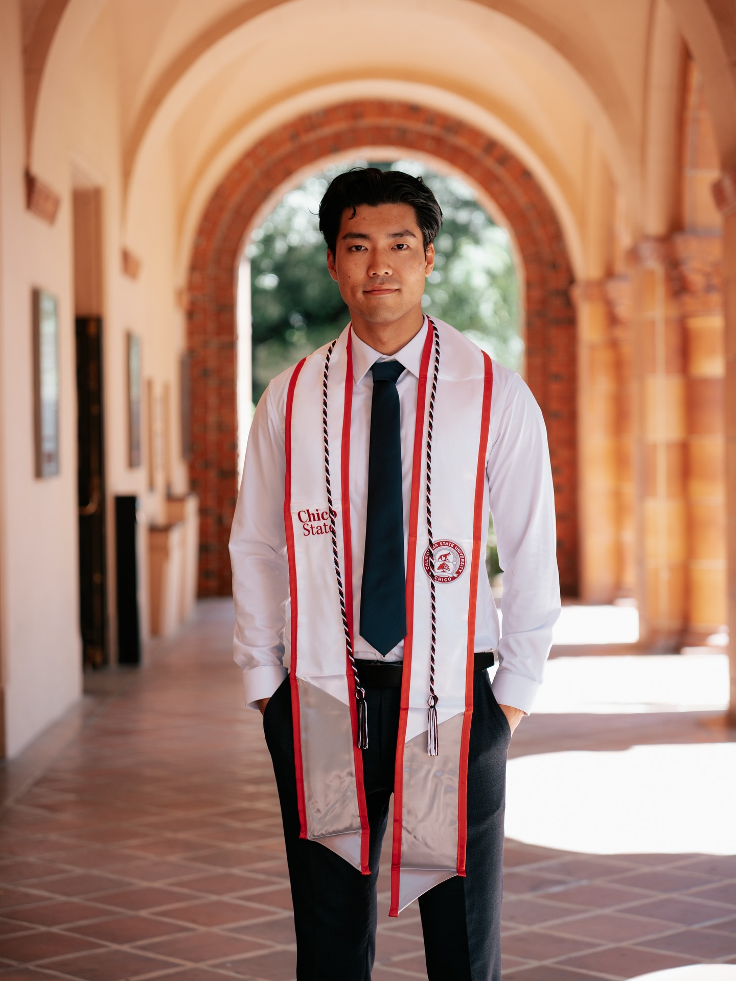 Graduate with hands in pockets in a long terracotta-arched walkway