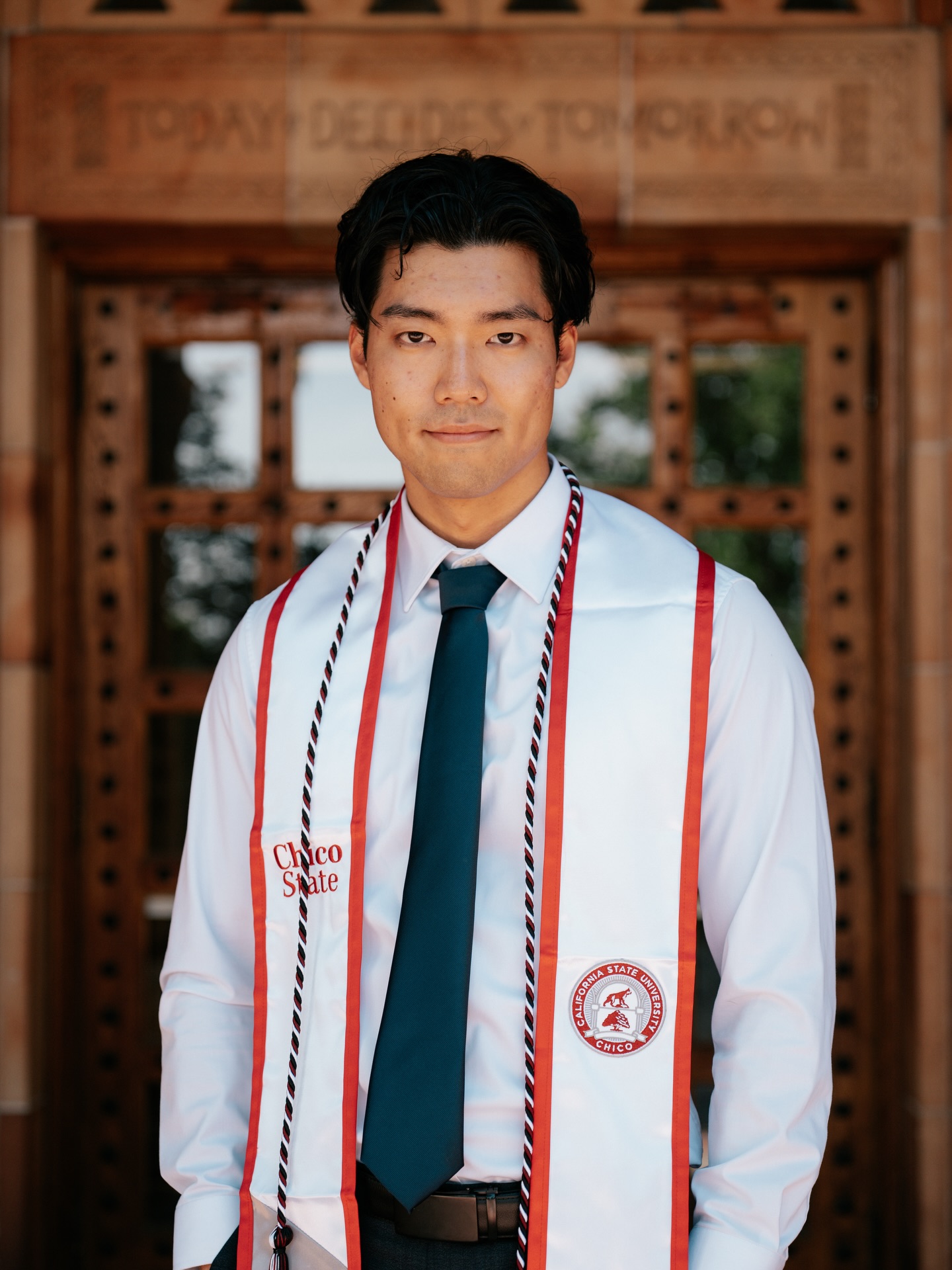Graduate in a white shirt, dark tie and red-and-white cords standing before carved wooden doors