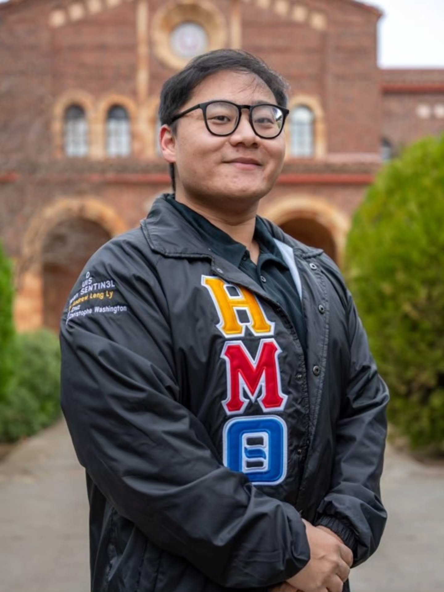 Chapter member in glasses and a black jacket with Greek letters, smiling in front of brick arches