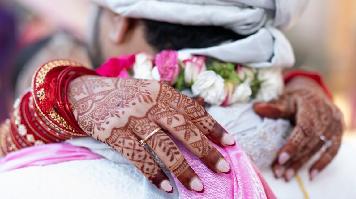 Henna-covered hands in red bangles resting on the groom’s shoulder beside a rose garland