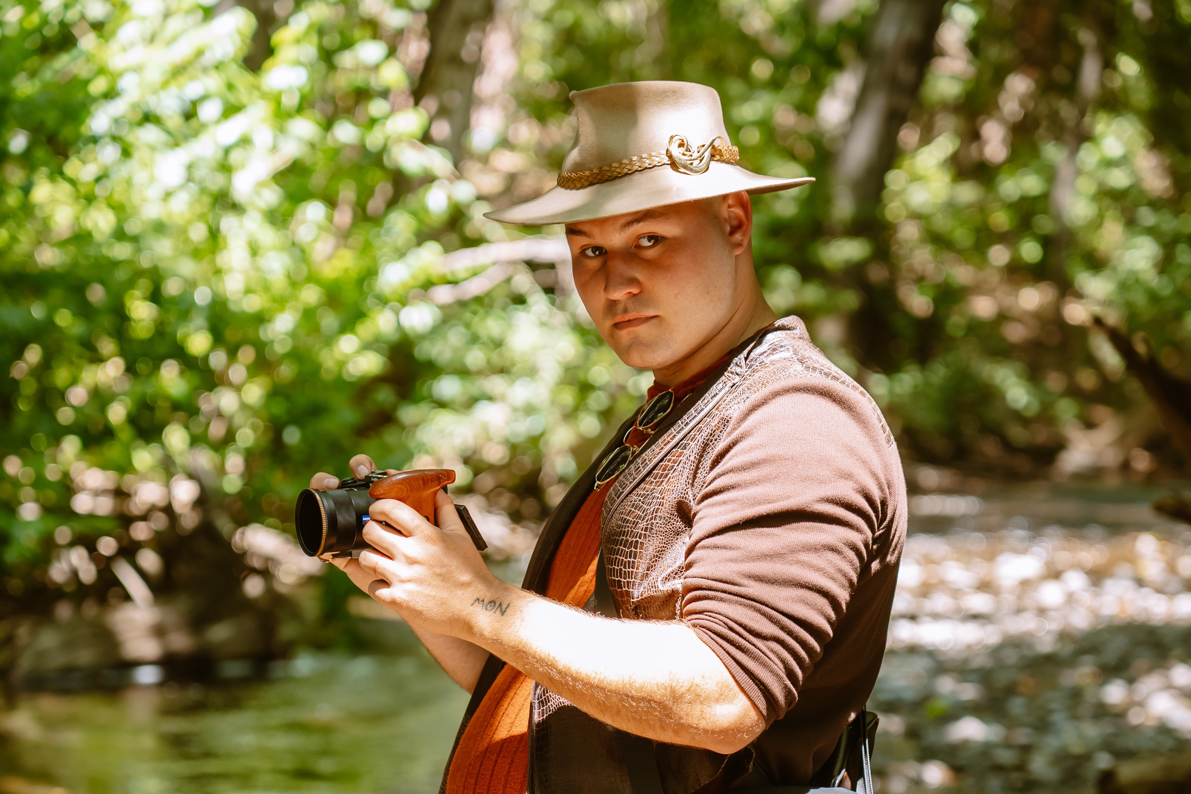 Crew member in a wide-brimmed hat holding a camera beside a creek
