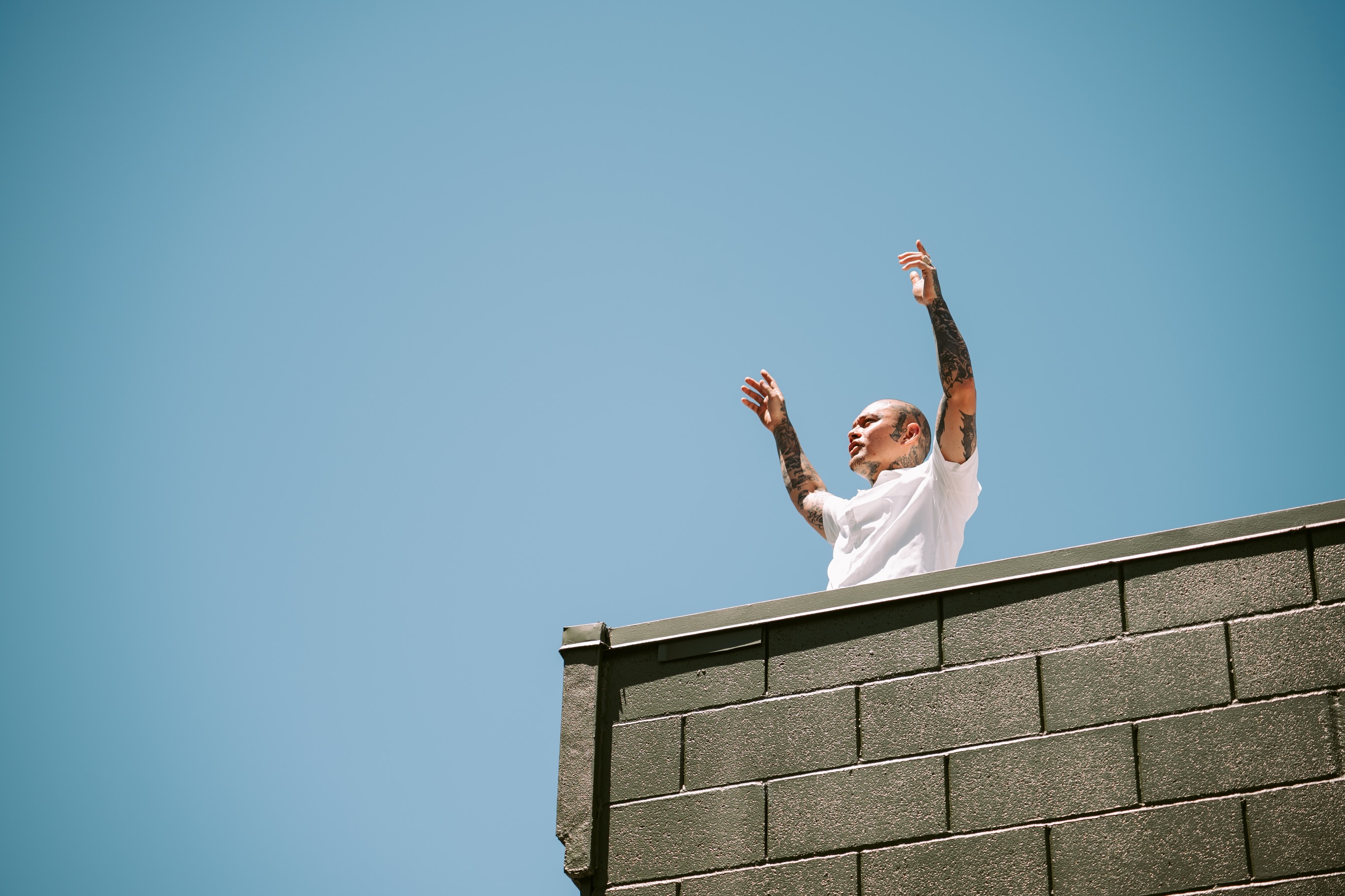 Tattooed subject in a white T-shirt raising both arms on a rooftop against a bright blue sky