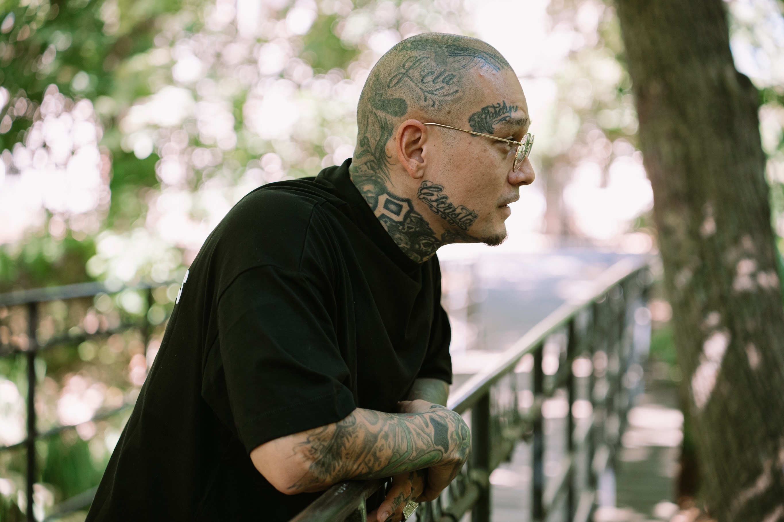 Tattooed subject in profile leaning on a metal railing under trees